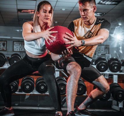 A man and woman perform a tandem workout with a medicine ball in a gym setting.