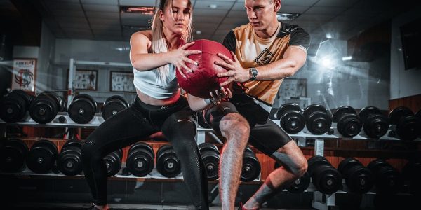 A man and woman perform a tandem workout with a medicine ball in a gym setting.