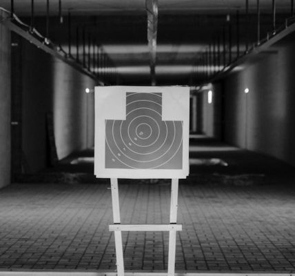 Black and white photo of a shooting range with a focus on a paper target in a dimly lit indoor setting.