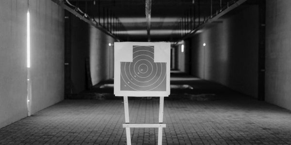 Black and white photo of a shooting range with a focus on a paper target in a dimly lit indoor setting.